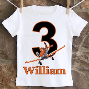 May include: White t-shirt with a black number 3 and a cartoon airplane in orange and white. The name "William" is printed in orange below the airplane.