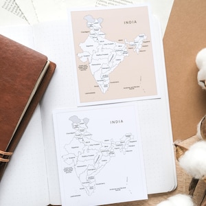 May include: Two square cards with a map of India, one in black and white, the other in beige. The cards are on a white dotted notebook, with a brown leather-bound journal and cotton buds in the background.
