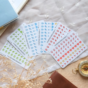 Puede incluir: Un conjunto de pegatinas Location Mini Icons en verde, azul y rojo, con formas como chinchetas, corazones y estrellas. Las pegatinas están dispuestas sobre una superficie clara, con una brújula y un cuaderno de cuero. Medidas en cm.