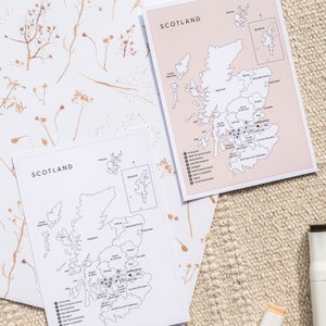 May include: Two black and white maps of Scotland with the regions labeled. The maps are on a beige surface with dried flowers.