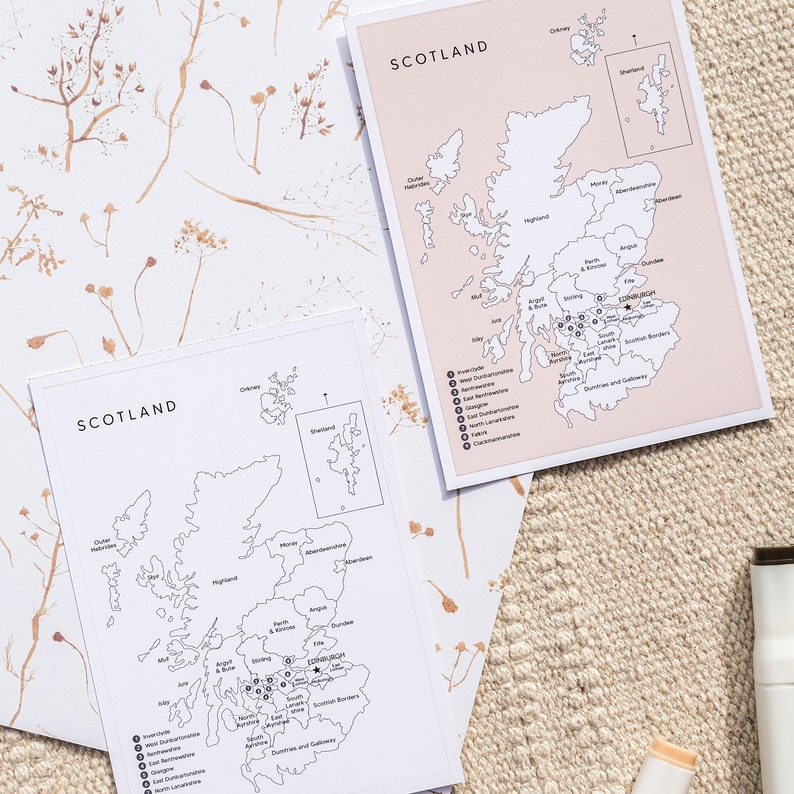 May include: Two black and white maps of Scotland with the regions labeled. The maps are on a beige surface with dried flowers.