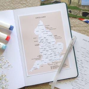 Könnte beinhalten: Ein offenes Notizbuch mit einer Karte von England auf hellrosa Hintergrund. Ein klarer Stift liegt auf der Seite. Das Wort "England" ist oben aufgedruckt. Drei farbige Marker sind links zu sehen.