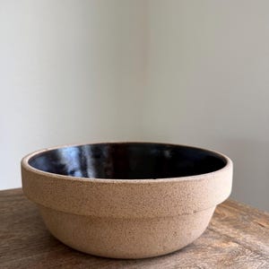 May include: A round ceramic bowl featuring a textured, speckled beige exterior and a smooth, glossy dark brown interior. The bowl has a distinct wide rim that encircles the upper portion, creating a clear separation between the base and the top edge.