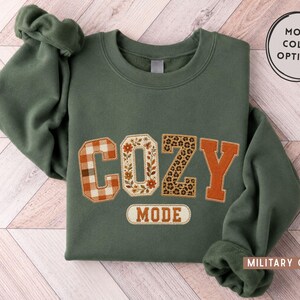 May include: A military green crewneck sweatshirt featuring the word COZY in large, multi-patterned applique letters, with a plaid C, floral O, leopard print Z, and solid orange Y. Below, the word MODE is embroidered in a rectangular patch.
