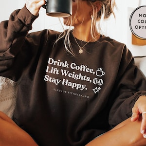 May include: A dark brown crewneck sweatshirt featuring white text that reads Drink Coffee, Lift Weights, Stay Happy. The design includes small white icons of a coffee mug, a dumbbell, and sparkles, with the text Vintage Fitness Club below.