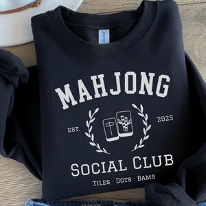 May include: Black sweatshirt with the text "MAHJONG SOCIAL CLUB" in white, along with the year 2025. The design includes a laurel wreath and images of Mahjong tiles. The sweatshirt also has the words "TILES DOTS BAMS".