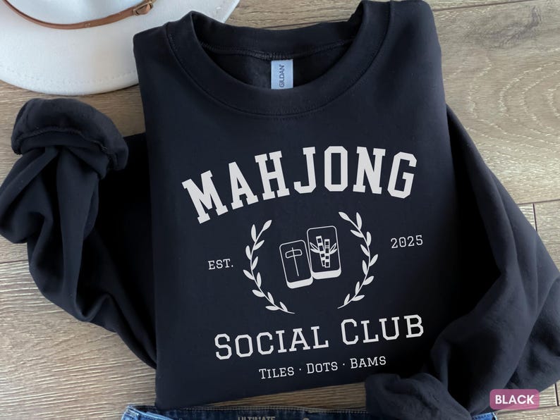 May include: Black sweatshirt with the text "MAHJONG SOCIAL CLUB" in white, along with the year 2025. The design includes a laurel wreath and images of Mahjong tiles. The sweatshirt also has the words "TILES DOTS BAMS".