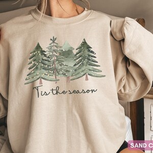 May include: A sand-coloured sweatshirt displaying a watercolour design of evergreen trees adorned with fairy lights. The words "Tis the season" are printed below the trees in a script font. A festive and comfortable garment.