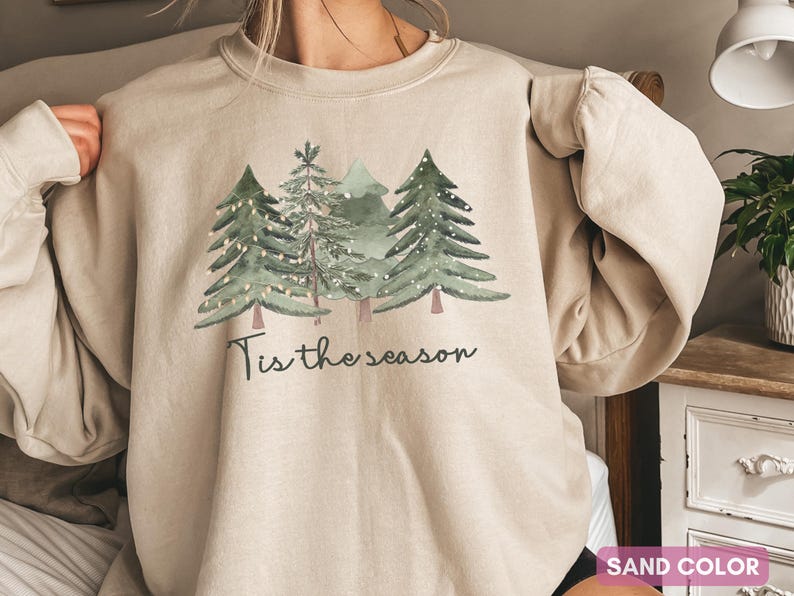 May include: A sand-coloured sweatshirt displaying a watercolour design of evergreen trees adorned with fairy lights. The words "Tis the season" are printed below the trees in a script font. A festive and comfortable garment.