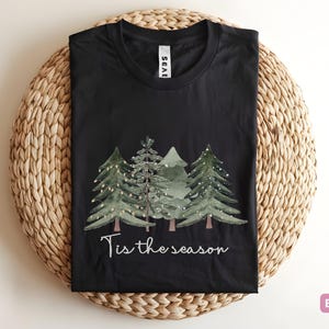 May include: Black t-shirt featuring a watercolour design of Christmas trees and the phrase "Tis the season". The trees are green with fairy lights. The t-shirt is displayed on a woven, circular placemat.