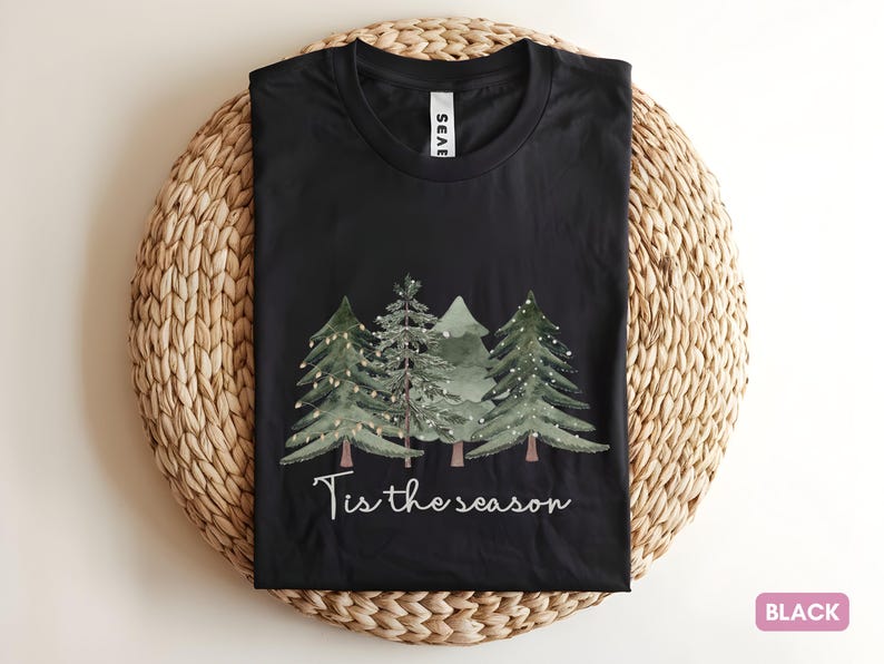 May include: Black t-shirt featuring a watercolour design of Christmas trees and the phrase "Tis the season". The trees are green with fairy lights. The t-shirt is displayed on a woven, circular placemat.