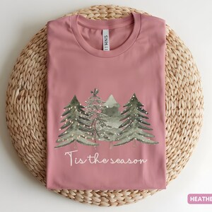 May include: Mauve t-shirt featuring a Christmas tree design and the text "Tis the season". The shirt is laid on a circular woven mat. The shirt's colour is described as heather mauve.