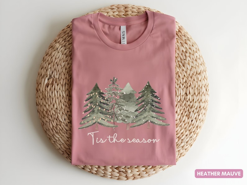 May include: Mauve t-shirt featuring a Christmas tree design and the text "Tis the season". The shirt is laid on a circular woven mat. The shirt's colour is described as heather mauve.
