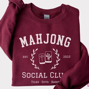 May include: Maroon sweatshirt with the text "MAHJONG SOCIAL CLUB" in white, along with the establishment year "2025". The design includes illustrations of Mahjong tiles and a laurel wreath. The bottom of the sweatshirt reads "TILES &bull; DOTS &bull; BAMS".