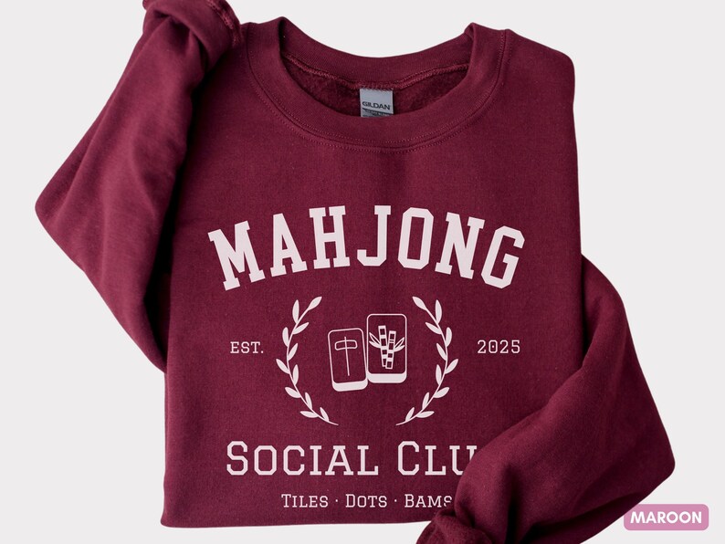 May include: Maroon sweatshirt with the text "MAHJONG SOCIAL CLUB" in white, along with the establishment year "2025". The design includes illustrations of Mahjong tiles and a laurel wreath. The bottom of the sweatshirt reads "TILES &bull; DOTS &bull; BAMS".