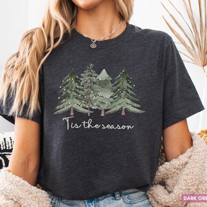 May include: Dark grey heather t-shirt with a watercolour design of three Christmas trees and fairy lights. The phrase "Tis the season" is written in a script font. A festive, cosy tee for the holiday season.