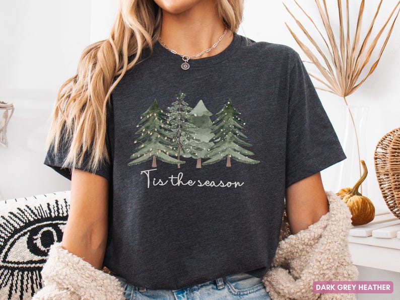 May include: Dark grey heather t-shirt with a watercolour design of three Christmas trees and fairy lights. The phrase "Tis the season" is written in a script font. A festive, cosy tee for the holiday season.