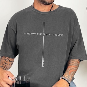 Puede incluir: Camiseta gris oscuro con el texto "THE WAY, THE TRUTH, THE LIFE" y una línea vertical, con "JOHN 14:6" debajo. La camiseta tiene mangas cortas y cuello redondo. La imagen también incluye un botón "MORE COLOR OPTIONS!"