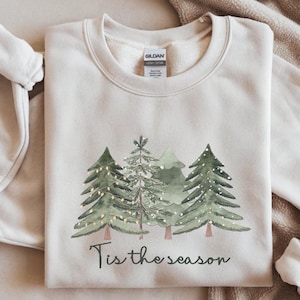 May include: A cream-coloured sweatshirt with a watercolour design of evergreen trees and fairy lights. The phrase "Tis the season" is printed below the trees. The sweatshirt is folded on a neutral-toned surface.