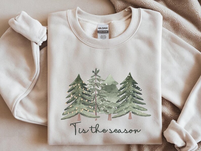 May include: A cream-coloured sweatshirt with a watercolour design of evergreen trees and fairy lights. The phrase "Tis the season" is printed below the trees. The sweatshirt is folded on a neutral-toned surface.