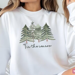May include: White sweatshirt with a watercolour design of evergreen trees and fairy lights. The phrase "Tis the season" is written below the trees. The sweatshirt is a crew neck style.