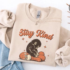 May include: Beige sweatshirt with the words "Stay Kind" in orange script and a graphic of a sad-looking monkey hugging a teddy bear. The sweatshirt has a crew neck and long sleeves. The design includes orange stars.