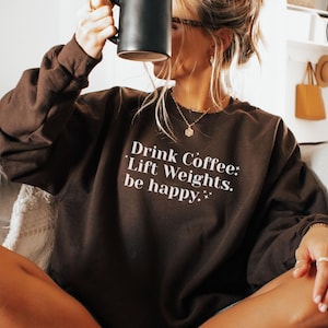 May include: A dark brown sweatshirt with the text "Drink Coffee: Lift Weights: be happy." in white. The person is holding a black mug. The sweatshirt is a relaxed fit and suitable for casual wear.