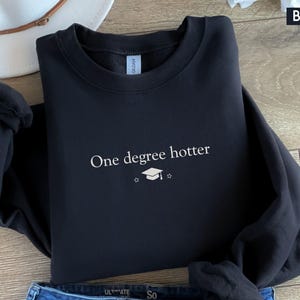 Pu&ograve; includere: Felpa nera con la scritta bianca "One degree hotter", un cappello di laurea e una stella. La parola "BLACK" &egrave; nell'angolo in alto a destra. Un cappello bianco e jeans blu sono parzialmente visibili. La felpa &egrave; piegata.