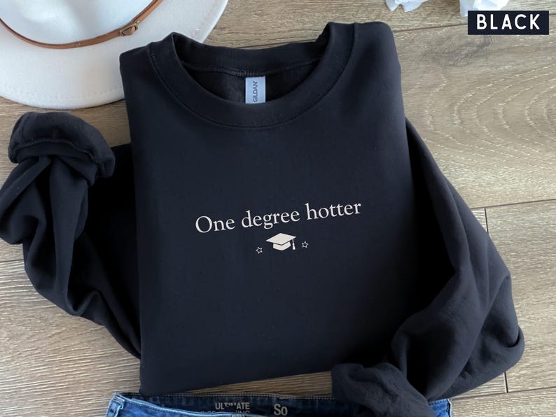Pu&ograve; includere: Felpa nera con la scritta bianca "One degree hotter", un cappello di laurea e una stella. La parola "BLACK" &egrave; nell'angolo in alto a destra. Un cappello bianco e jeans blu sono parzialmente visibili. La felpa &egrave; piegata.