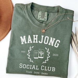 May include: An olive green t-shirt with the word "MAHJONG" in white block letters. Below the word is "SOCIAL CLUB" and the text "TILES DOTS BAMS". A gold necklace and a brown hat are also in the image.