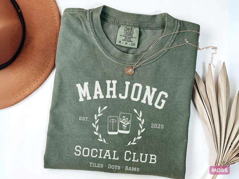 May include: An olive green t-shirt with the word "MAHJONG" in white block letters. Below the word is "SOCIAL CLUB" and the text "TILES DOTS BAMS". A gold necklace and a brown hat are also in the image.