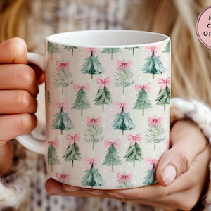 May include: White ceramic mug featuring a repeating pattern of watercolor Christmas trees and bows in shades of green and pink on a light beige background. The mug has a white handle and is held by a person.
