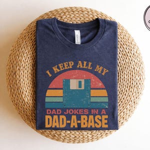 May include: Navy blue t-shirt with the text "I KEEP ALL MY DAD JOKES IN A DAD-A-BASE" in orange and white. The graphic features a retro sunset design with a floppy disk. The shirt is folded on a woven surface.