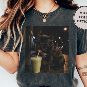 May include: A charcoal gray t-shirt featuring a graphic print of a black French bulldog wearing sunglasses and drinking from a clear plastic cup with a straw. The shirt includes a circular badge in the top right corner with the text MORE COLOR OPTIONS!