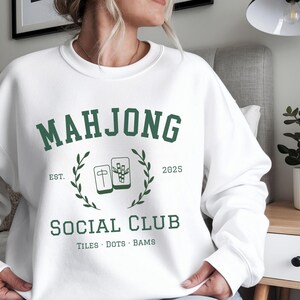 May include: White sweatshirt with a green "MAHJONG SOCIAL CLUB" graphic. The design includes the text "EST. 2025" and illustrations of Mahjong tiles. The sweatshirt is a casual garment, suitable for everyday wear.