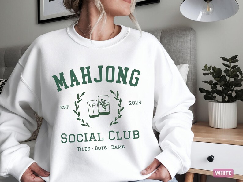 May include: White sweatshirt with a green "MAHJONG SOCIAL CLUB" graphic. The design includes the text "EST. 2025" and illustrations of Mahjong tiles. The sweatshirt is a casual garment, suitable for everyday wear.