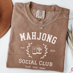 May include: Espresso-colored t-shirt with the text "MAHJONG SOCIAL CLUB" in white. The shirt features a graphic of mahjong tiles and the year 2025. A brown hat and silver necklace with a pendant are also visible.