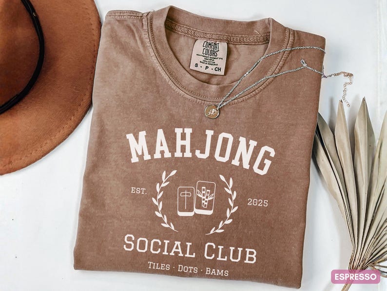 May include: Espresso-colored t-shirt with the text "MAHJONG SOCIAL CLUB" in white. The shirt features a graphic of mahjong tiles and the year 2025. A brown hat and silver necklace with a pendant are also visible.