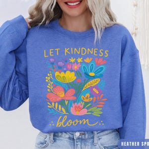 May include: Heather sport royal blue sweatshirt with a floral design. The design features colorful flowers and the words "Let Kindness Bloom" in yellow and white. The sweatshirt is a crew neck style.