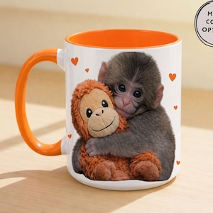 May include: White ceramic mug with an orange interior and handle. The mug features a print of a baby monkey hugging a stuffed orangutan. Small red hearts are scattered around the image. The mug has the text "MORE COLOR OPTIONS!"