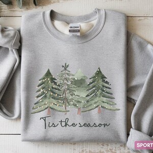 May include: Sporty grey sweatshirt featuring a watercolour design of Christmas trees and the phrase "Tis the season" in black script. The sweatshirt is made of a soft material, ideal for the festive season.