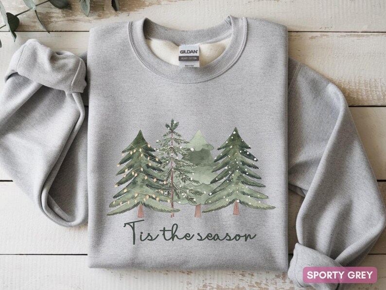 May include: Sporty grey sweatshirt featuring a watercolour design of Christmas trees and the phrase "Tis the season" in black script. The sweatshirt is made of a soft material, ideal for the festive season.