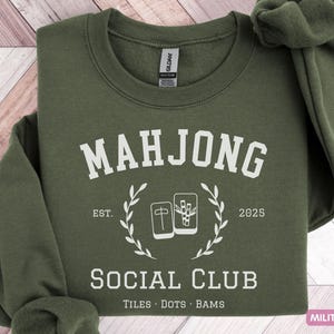 May include: Military green sweatshirt with the text "MAHJONG SOCIAL CLUB" in white. The design includes a laurel wreath and Mahjong tiles. The sweatshirt is a crew neck style.