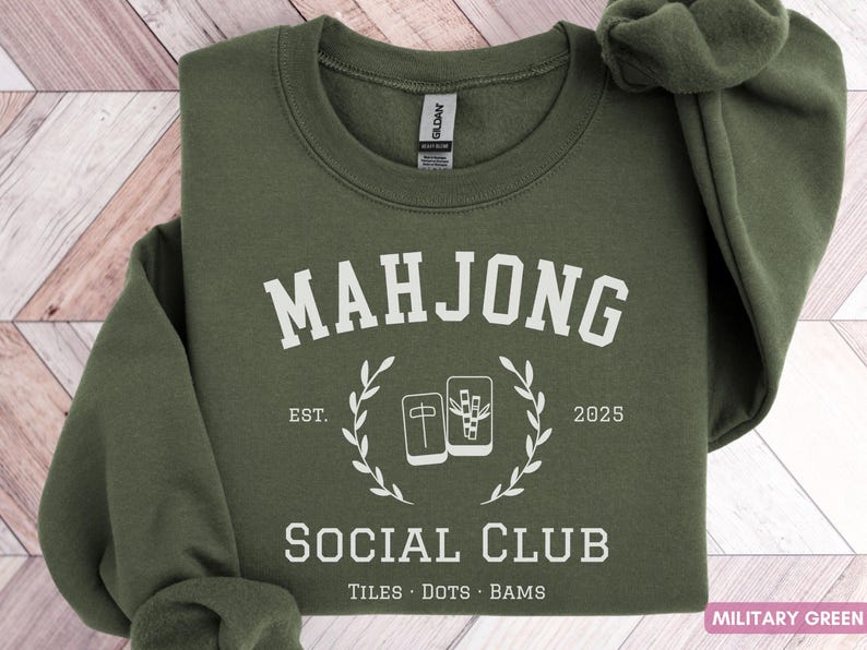 May include: Military green sweatshirt with the text "MAHJONG SOCIAL CLUB" in white. The design includes a laurel wreath and Mahjong tiles. The sweatshirt is a crew neck style.