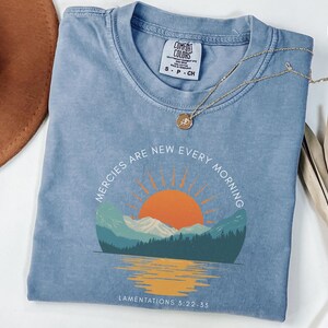 Puede incluir: Camiseta azul claro con el texto "Mercies are new every morning" que rodea un gráfico de un amanecer sobre montañas y agua. Un collar dorado descansa sobre la camisa. La camisa tiene una etiqueta Comfort Colors.