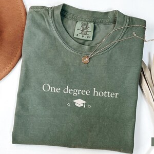 Pu&ograve; includere: Una maglietta verde muschio con la scritta "One degree hotter" e una grafica di cappello di laurea. Una collana d'oro con un ciondolo poggia sulla maglietta. Un cappello marrone e una pianta secca sono sullo sfondo.