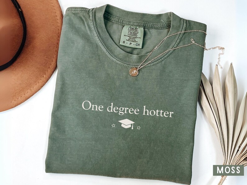 Pu&ograve; includere: Una maglietta verde muschio con la scritta "One degree hotter" e una grafica di cappello di laurea. Una collana d'oro con un ciondolo poggia sulla maglietta. Un cappello marrone e una pianta secca sono sullo sfondo.
