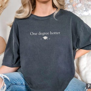 Pu&ograve; includere: T-shirt grigio scuro con la scritta bianca "One degree hotter", una stella e un cappello di laurea. La maglietta ha maniche corte e una vestibilit&agrave; ampia. La parola "PEPPER" &egrave; stampata in basso a destra.