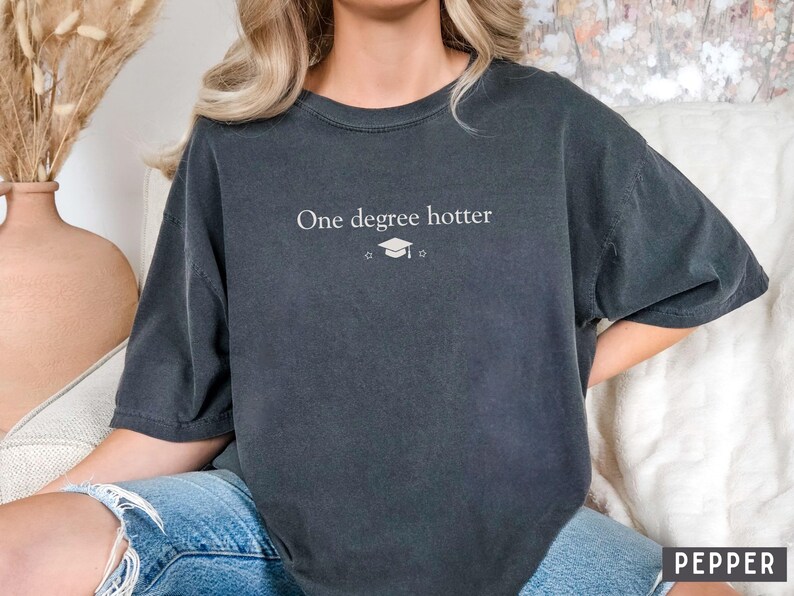 Pu&ograve; includere: T-shirt grigio scuro con la scritta bianca "One degree hotter", una stella e un cappello di laurea. La maglietta ha maniche corte e una vestibilit&agrave; ampia. La parola "PEPPER" &egrave; stampata in basso a destra.