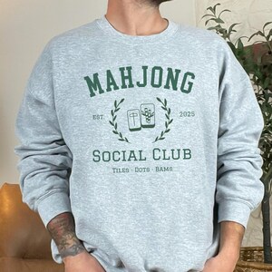 May include: A light gray sweatshirt with the words "MAHJONG SOCIAL CLUB" in green, along with the year "2025" and the words "TILES DOTS BAMS". The sweatshirt has a crew neck and long sleeves.